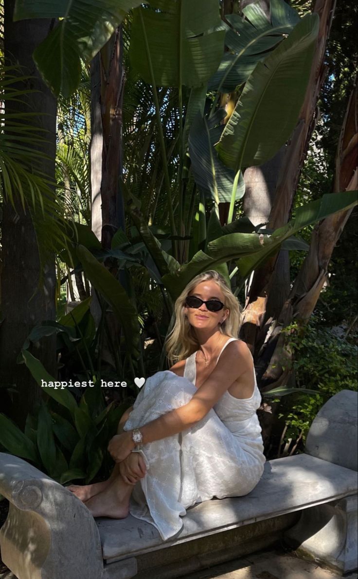 a woman sitting on top of a stone bench in front of palm trees and greenery