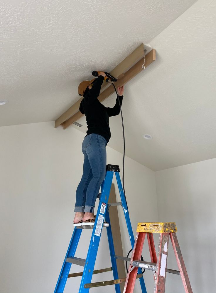 a woman standing on top of a ladder working on a beam in a room with white walls