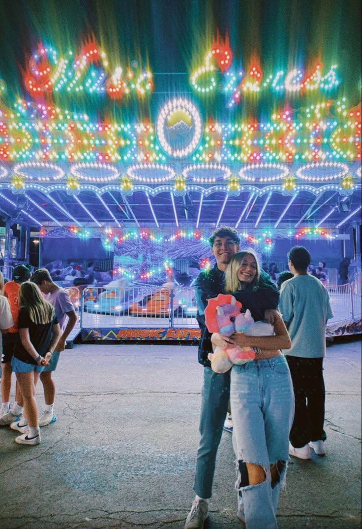 two people standing in front of a carnival ride at night with lights on the marquee