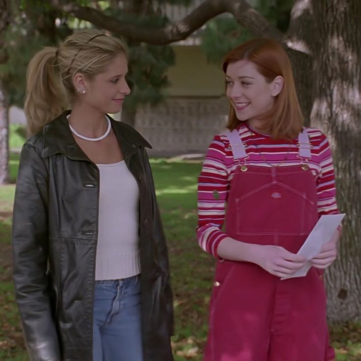 two women standing next to each other in front of a tree and one is holding papers