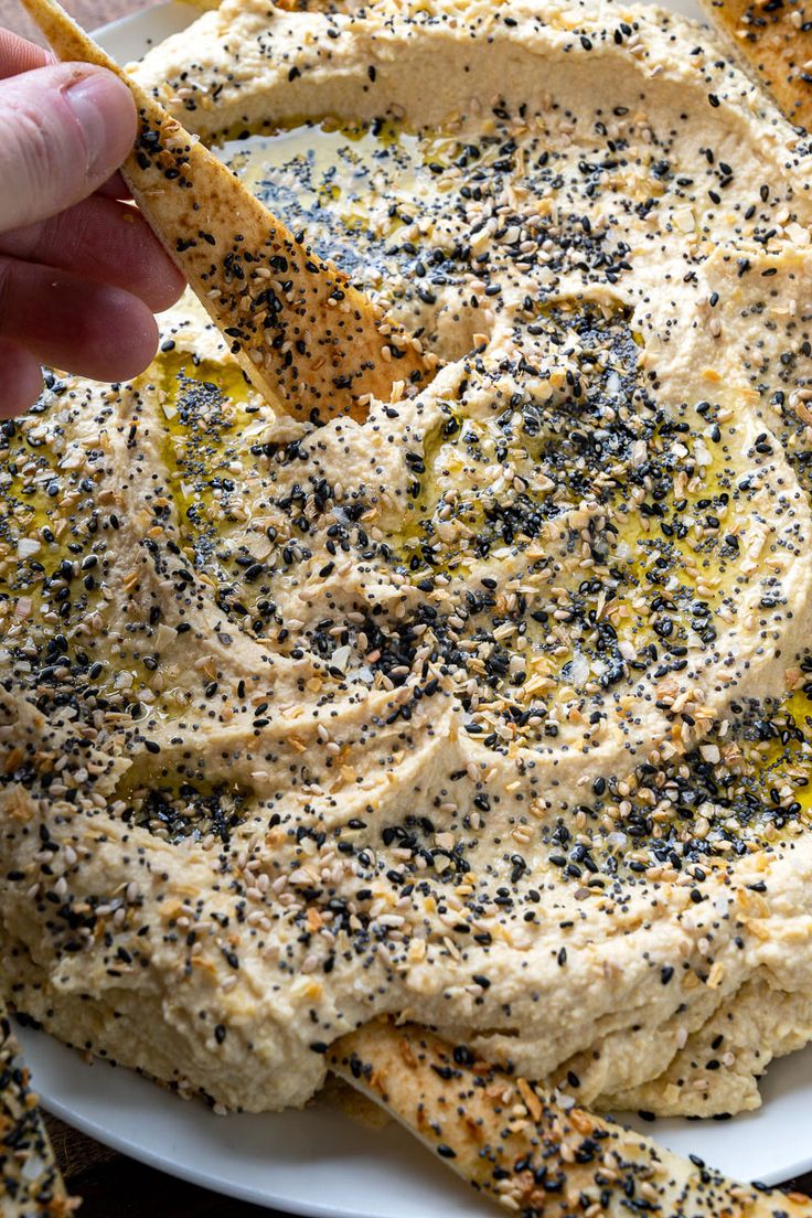 a hand dipping hummus into a white bowl filled with mustard and black sesame seeds