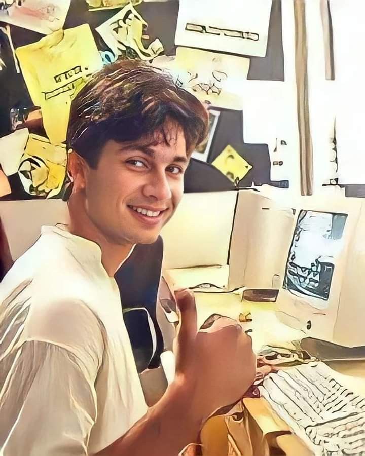 a young man sitting in front of a desk with a computer on it and lots of sticky notes all over the wall behind him