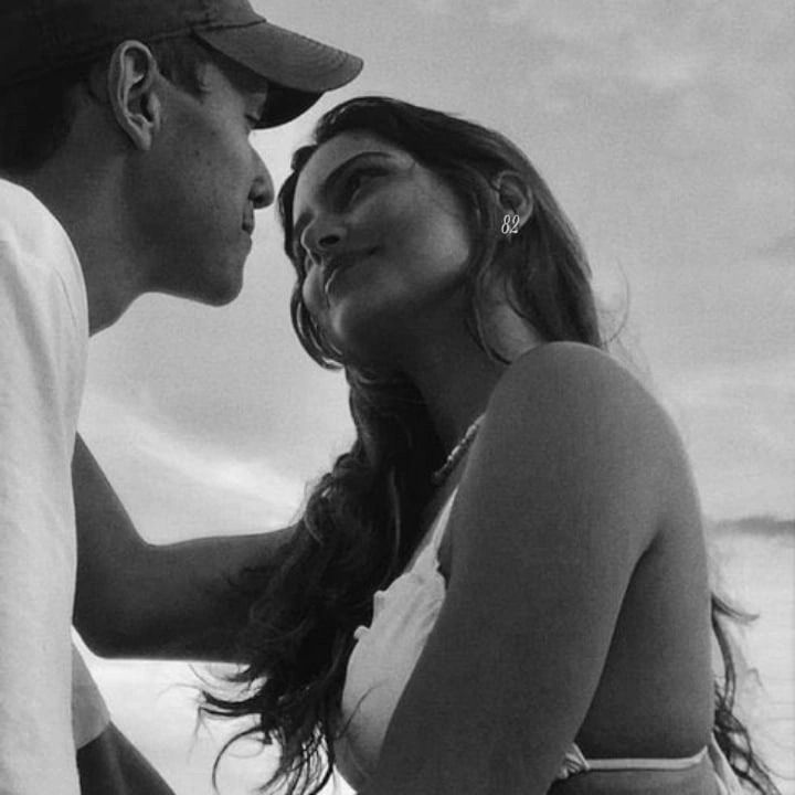 a man and woman sitting next to each other on the beach, one is kissing the other's forehead