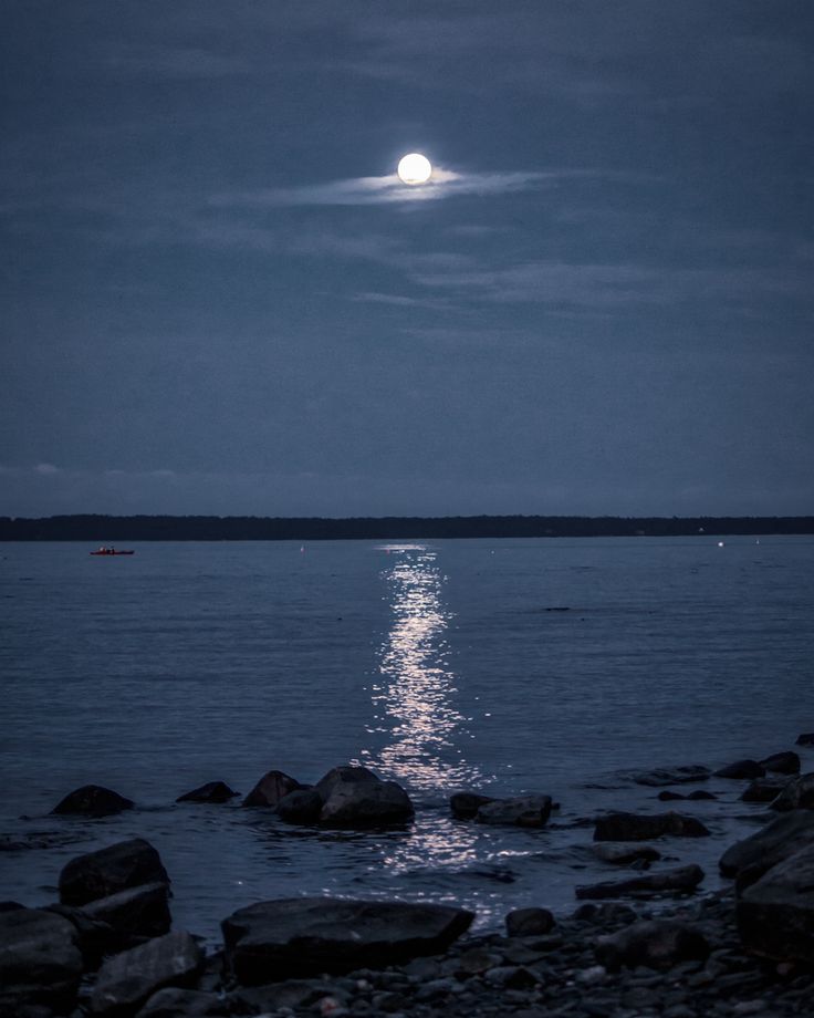 the moon shines brightly in the night sky over water with rocks on the shore