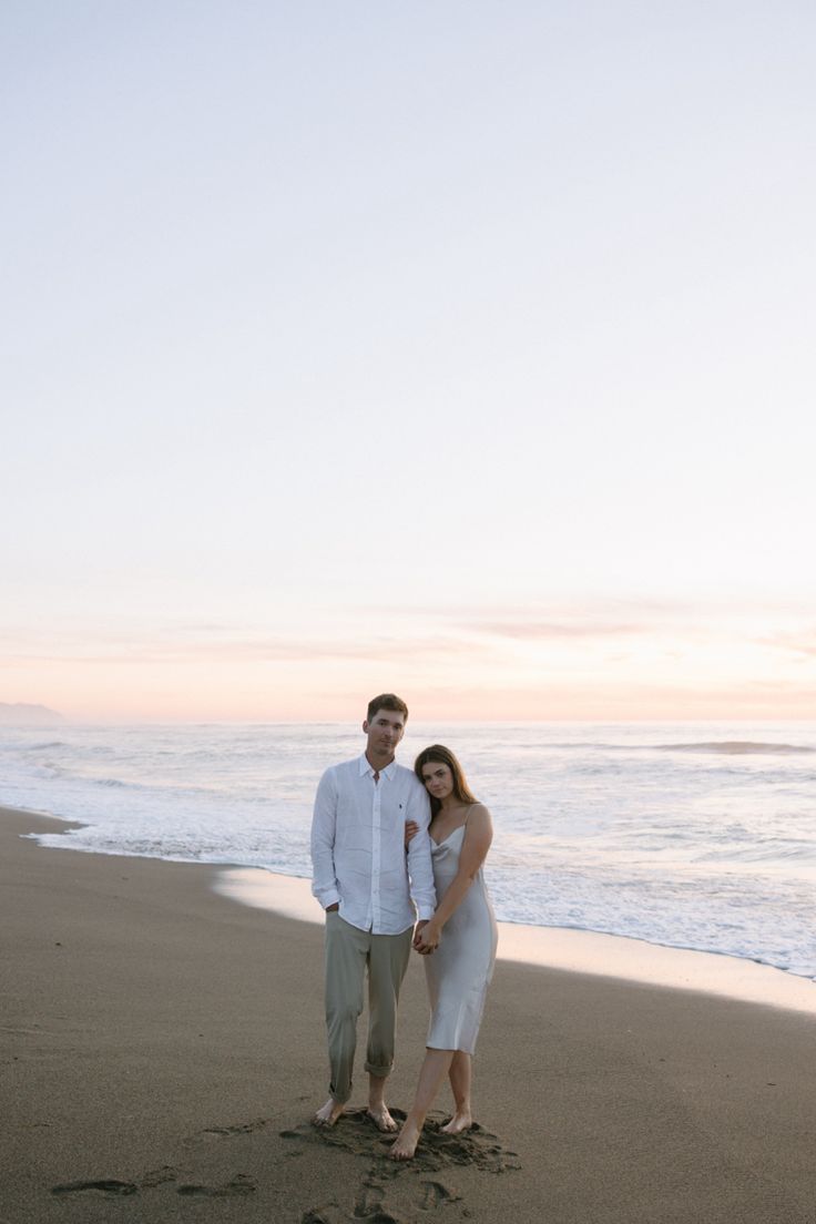 a man and woman standing on top of a sandy beach next to the ocean at sunset