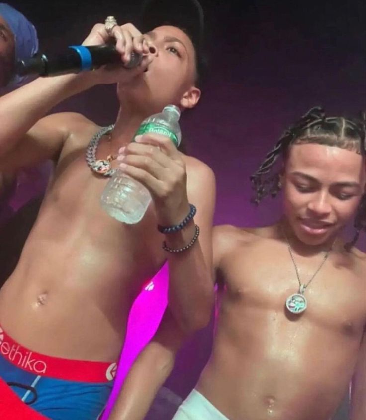 two men with no shirts on singing into microphones while another man holds a bottle in his hand