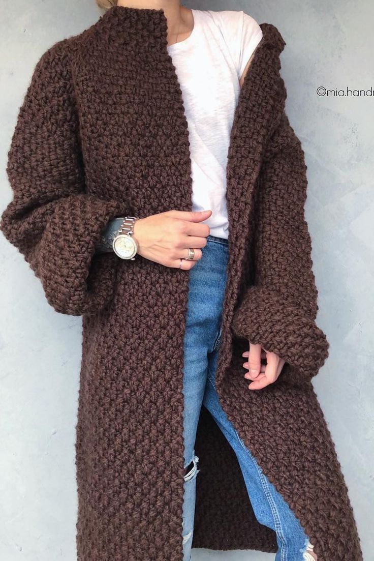 a woman wearing a long brown cardigan sweater and jeans, standing against a wall