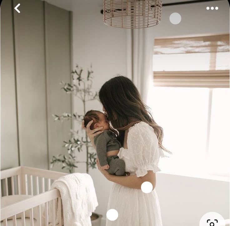 a woman in a white dress holding a baby next to a crib and window