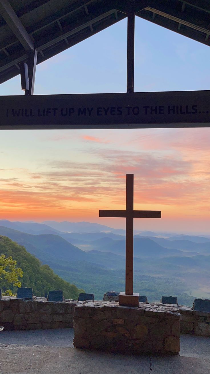 a wooden cross sitting on top of a stone slab under a roof next to a sign that says, will lift my eyes to the hills