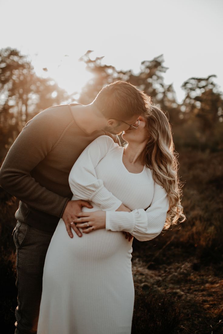 a pregnant couple kissing in the woods at sunset