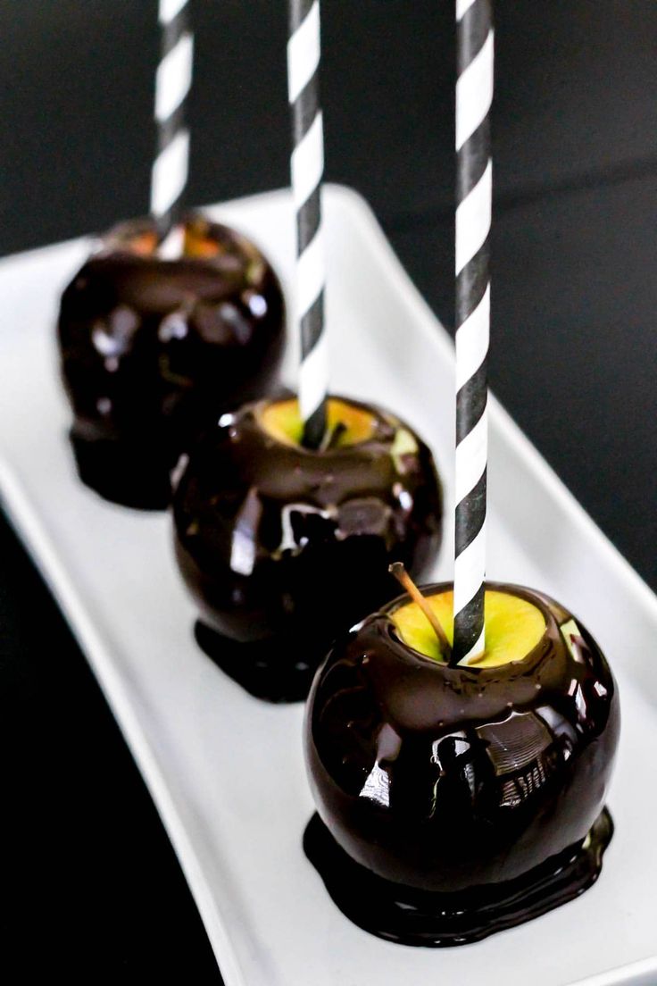 three chocolate covered apples on a white plate with black and white striped sticks sticking out of them