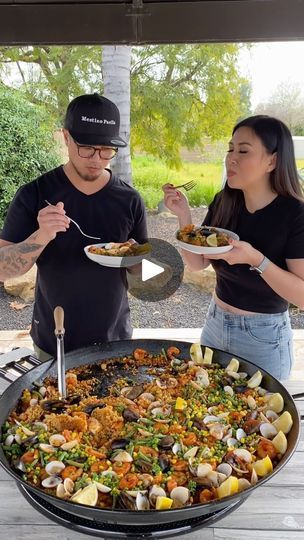 two people standing in front of a large pan filled with food