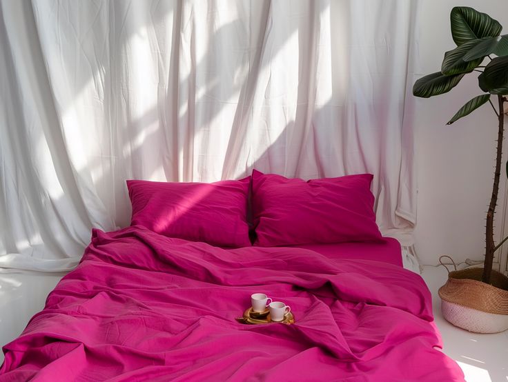 a bed with pink sheets and pillows in front of a white curtained wall, next to a potted plant