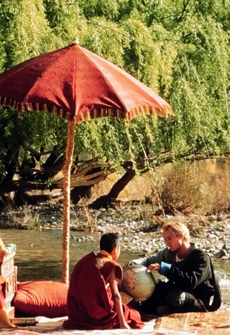 three people sitting on the ground under an umbrella next to a body of water with trees in the background