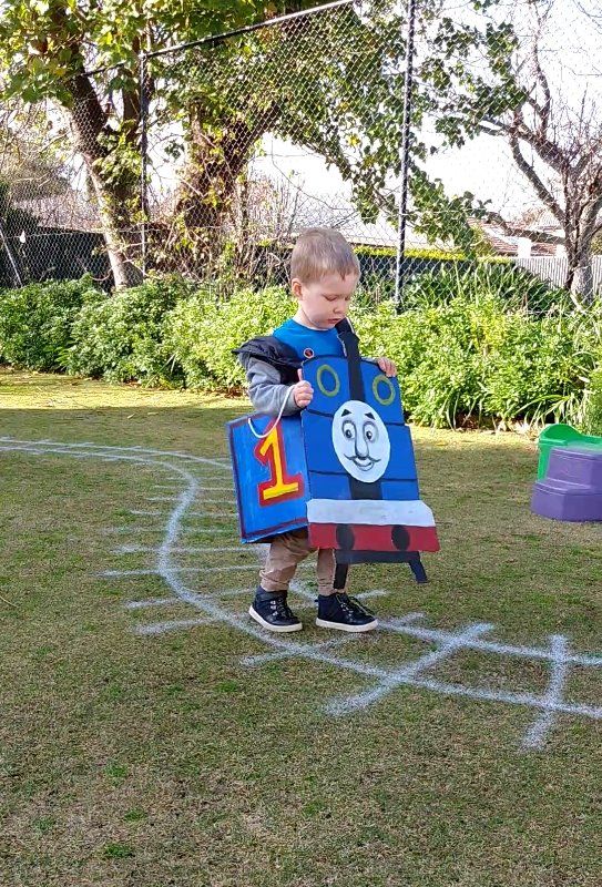 a little boy standing on top of a field holding a blue train shaped cardboard box