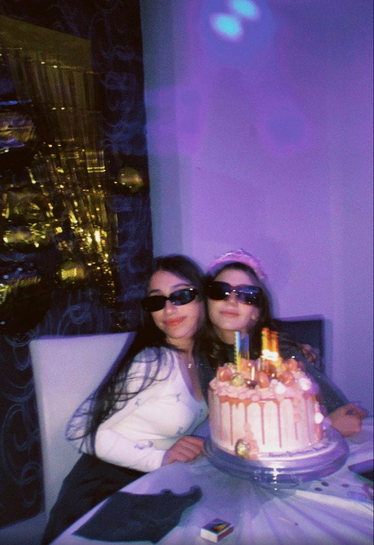 two women are sitting at a table with a cake in front of them and one is wearing sunglasses