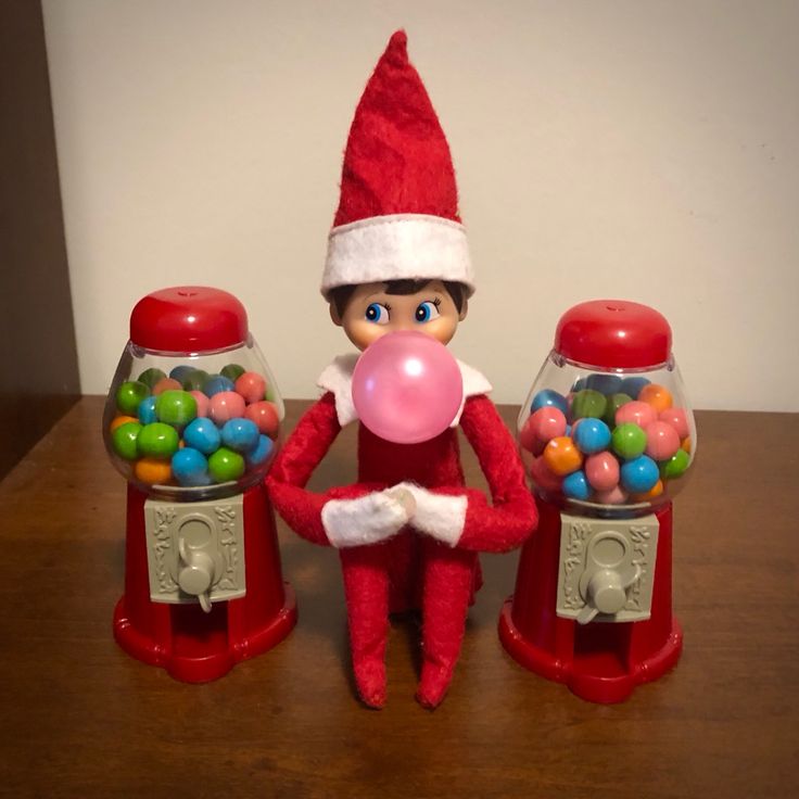 the elf is sitting next to two gumballs machines and holding a pink bubble ball