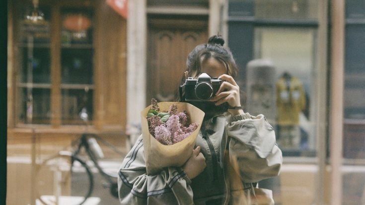 a woman taking a photo with her camera in front of a store window holding a bouquet of flowers