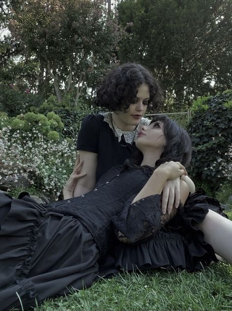 two women laying on the grass in front of some bushes and trees, one is kissing the other's head