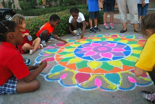 children are sitting on the ground and drawing with colored chalk in front of an adult