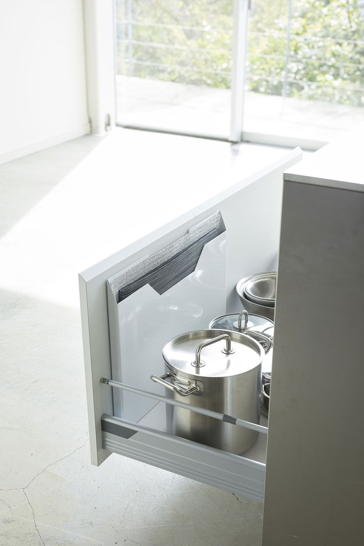 an open refrigerator door with pots and pans in the bottom drawer, next to a window