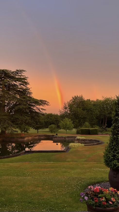 a rainbow is seen in the distance over a pond and garden at sunset or dawn