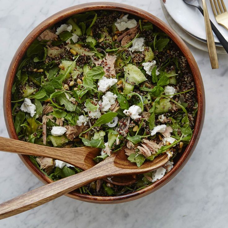 a salad in a bowl with two wooden spoons next to it on a marble table