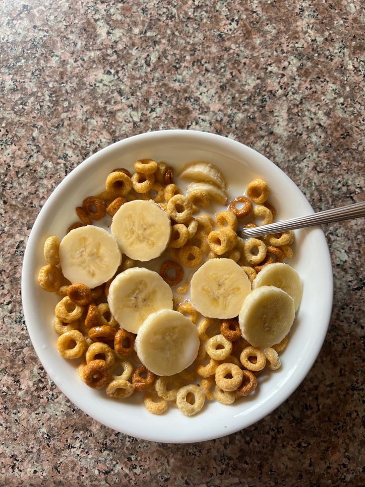 a bowl filled with cereal and bananas on top of a granite countertop next to a spoon