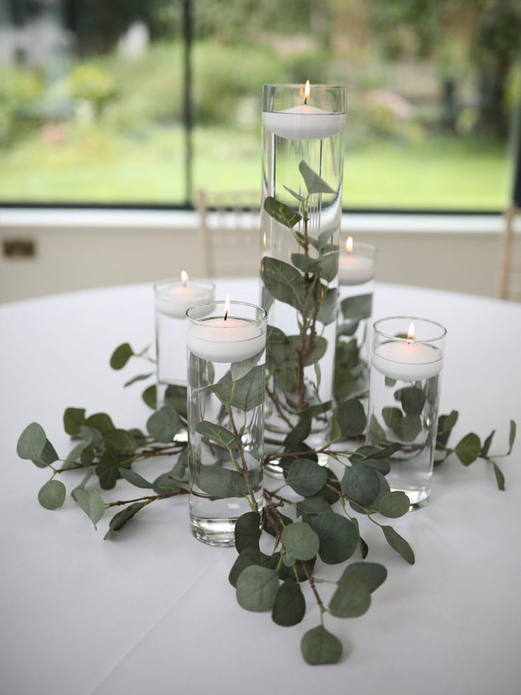 candles and greenery are arranged in glass vases on a white tablecloth with windows behind them