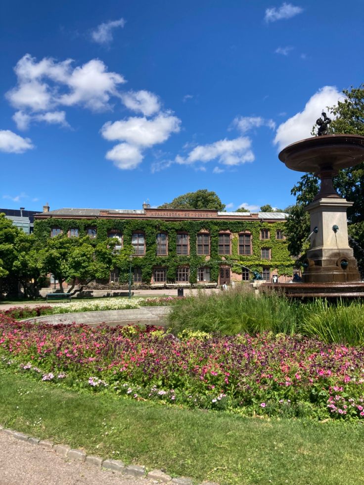 a fountain in the middle of a garden with flowers around it and a building in the background