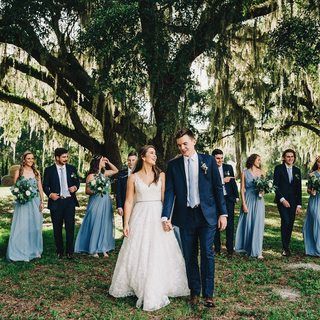 a bride and groom walking in front of their bridal party under the shade of a large tree