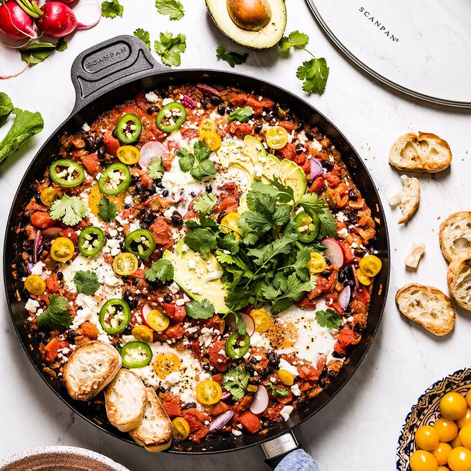 a skillet filled with vegetables and cheese on top of a white table next to bread