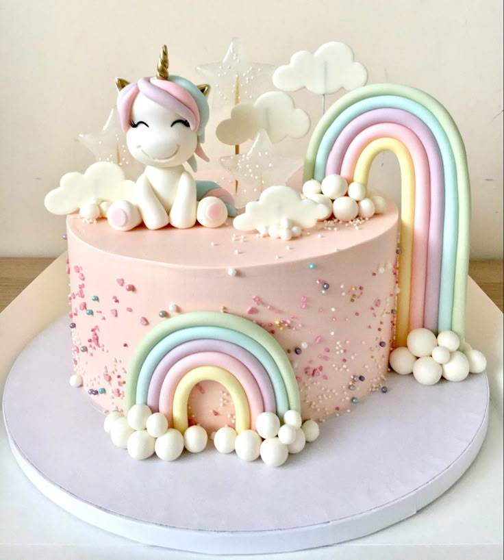 there is a pink cake with rainbows and clouds on the top that has a unicorn sitting on it