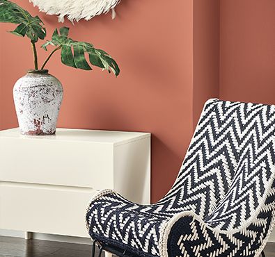 a chair with a plant in it next to a white dresser and pink wall paint