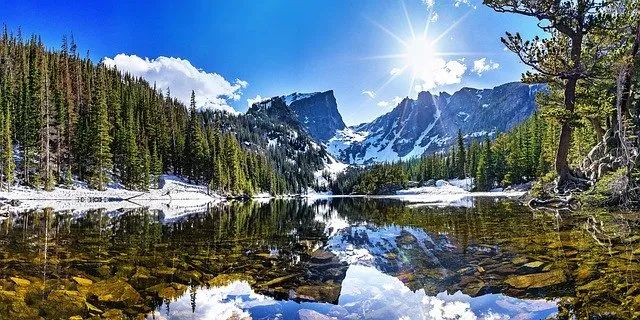 the sun shines brightly over a mountain lake surrounded by pine trees and snow covered mountains