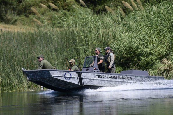 a group of men riding on the back of a boat in water next to tall grass