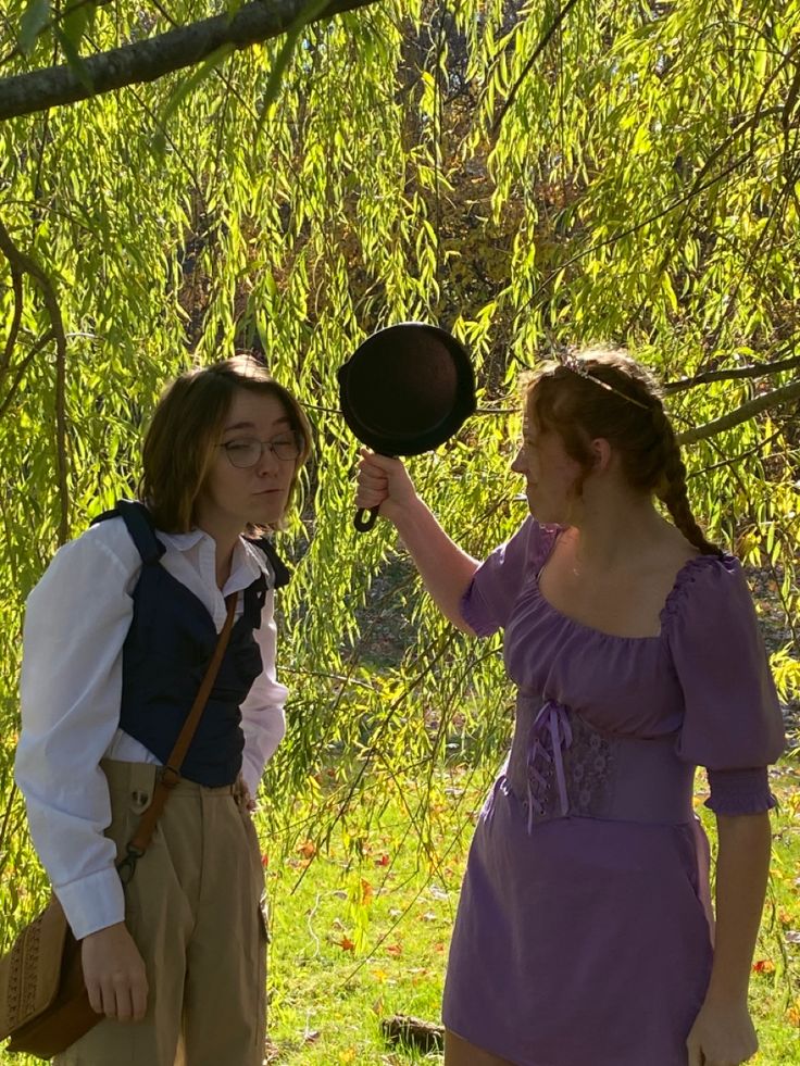 two women dressed in period clothing are standing under a tree and one is holding a frying pan