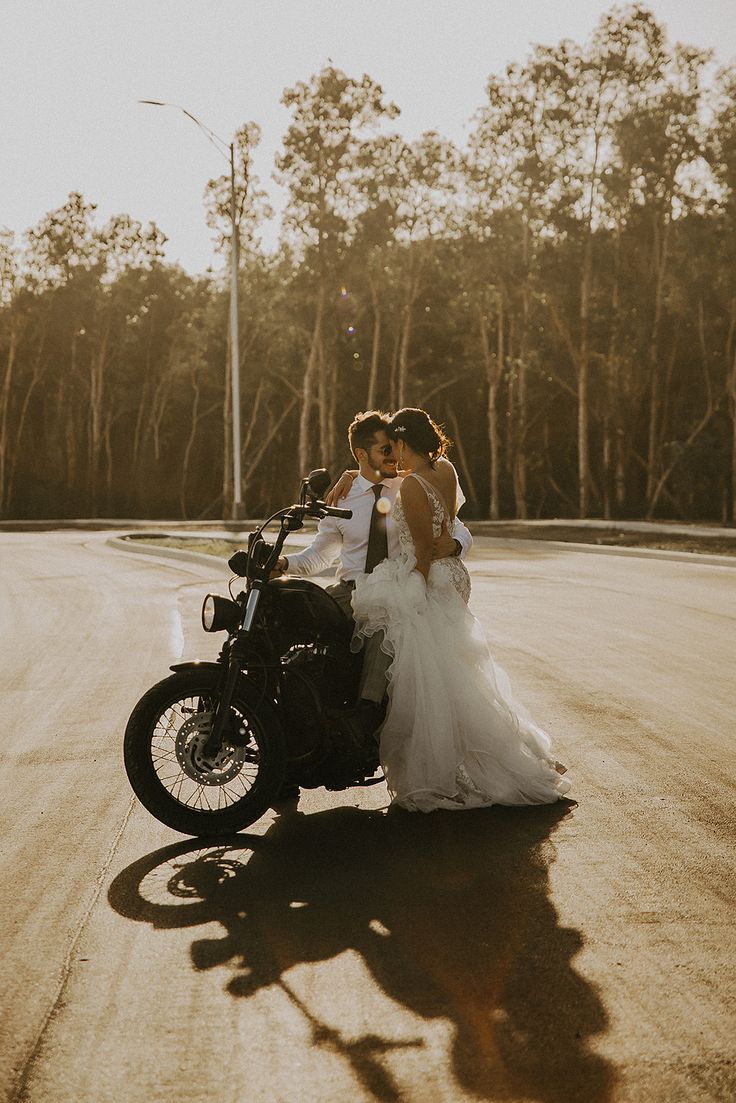 a bride and groom are standing next to a motorcycle on the road with trees in the background