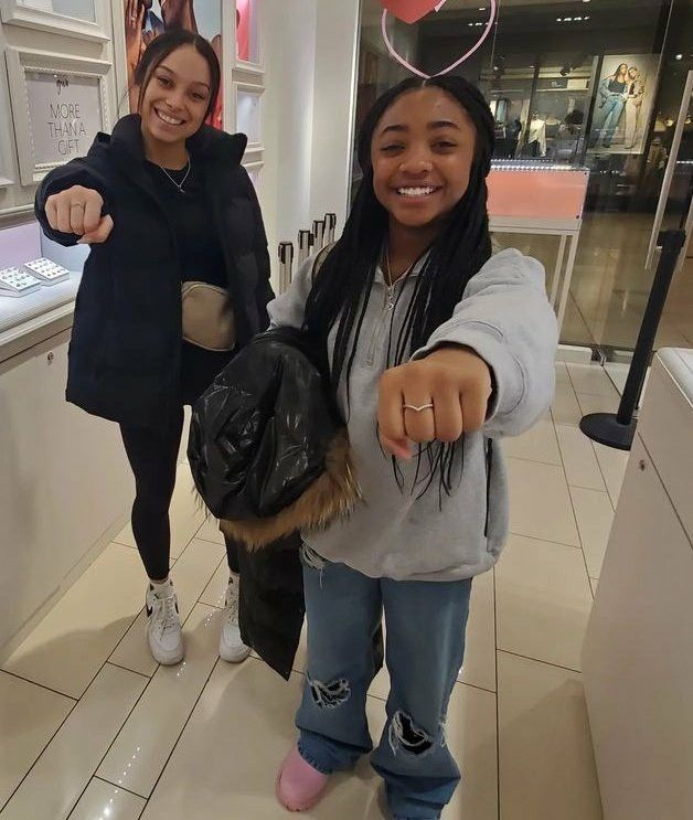 two girls are pointing at the camera in a shopping mall, one is wearing a backpack and the other has a hat on her head