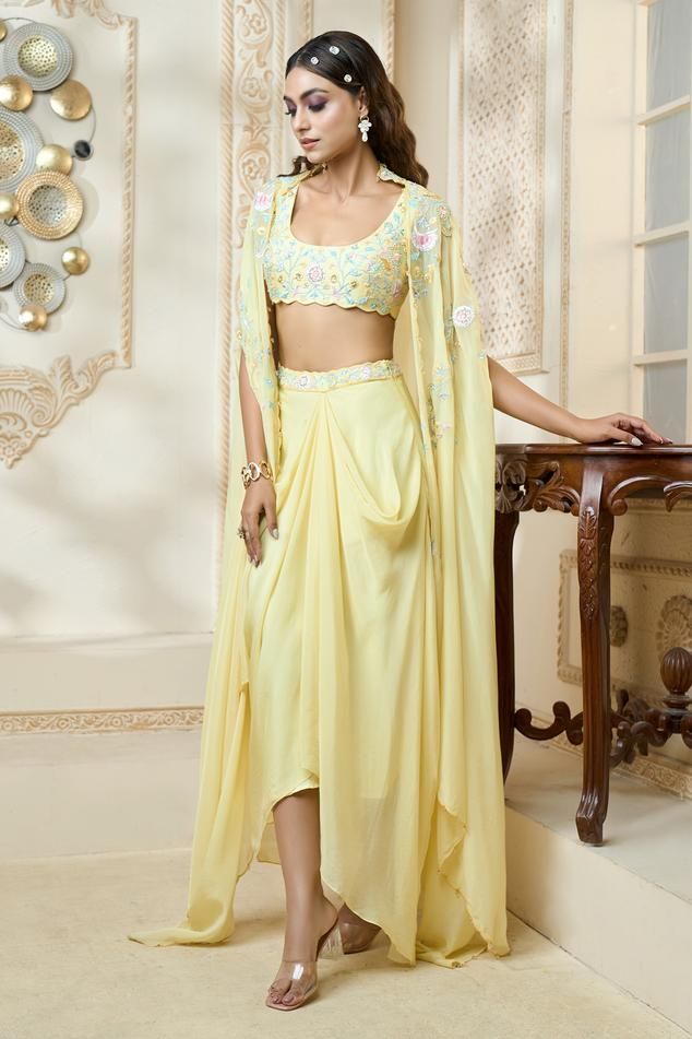 Buttercup draped skirt with floral embroidered waistband. Comes with floral embroidered bustier and flowy cape. 
Components: 3
Pattern: Embroidered
Type Of Work: Floral
Neckline: Bustier: Scoop
Sleeve Type: Bustier: Sleeveless, Cape: Draped
Fabric: Bustier: Dupion Silk, Cape and Skirt: Viscose Organza
Color: Yellow
Other Details: 
Scalloped hem bustier and cape
Closure: Skirt and Bustier: Side zip
Occasion: Mehendi and Haldi - Aza Fashions Cape Women, Embroidered Bustier, Cape For Women, Silk Cape, Haldi Outfit, Long Cape, Floral Bustier, Draped Skirt, Dupion Silk