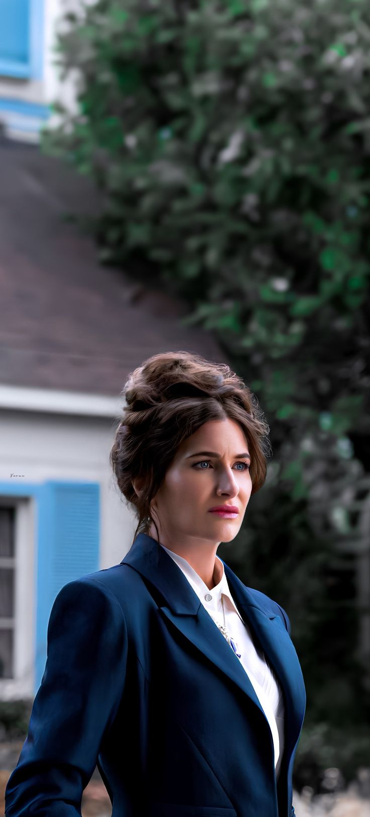 a woman standing in front of a house wearing a blue suit and white shirt with her hands on her hips