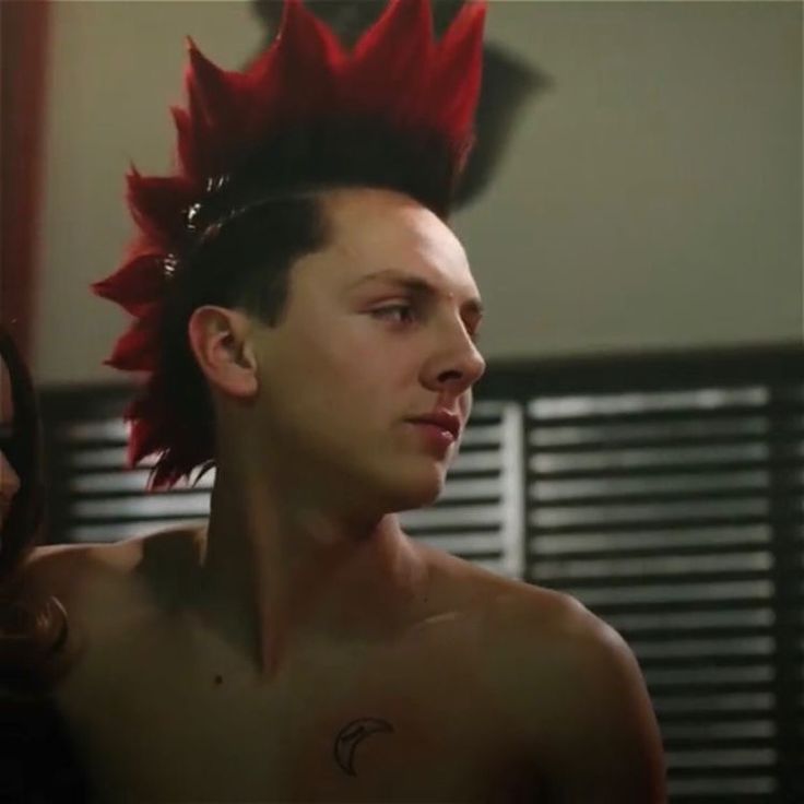 a shirtless man with red mohawks on his head and another woman in the background