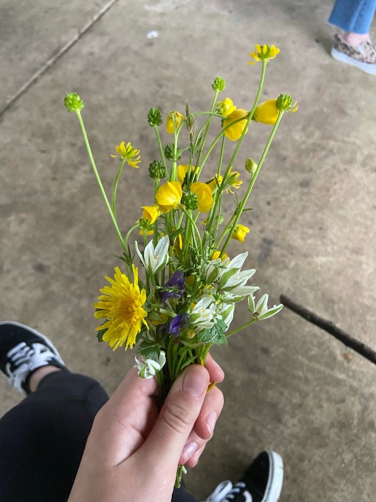 a person holding a bunch of flowers in their hand on the sidewalk with someone's feet