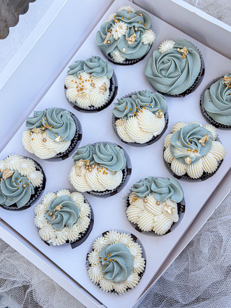 twelve cupcakes with blue frosting and gold sprinkles in a white box