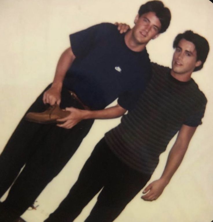 two men standing next to each other in front of a white wall with one man holding his hat