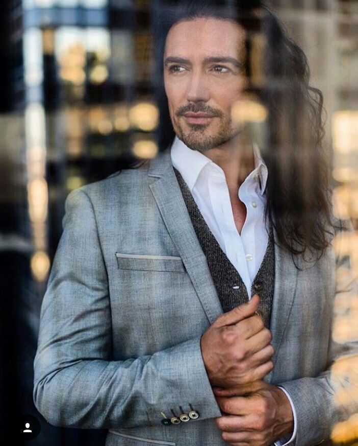 a man with long hair wearing a gray suit and white shirt standing in front of a window