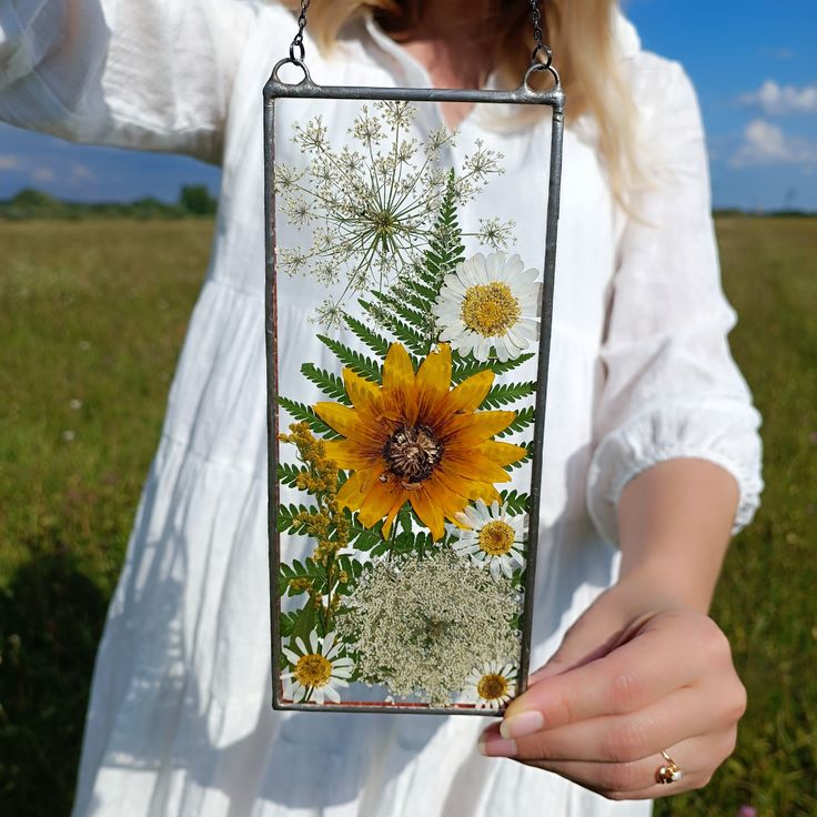 a woman holding up a glass block with flowers on it in the middle of a field