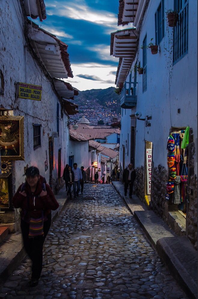 a narrow cobblestone street with shops and people walking on the sidewalk at dusk