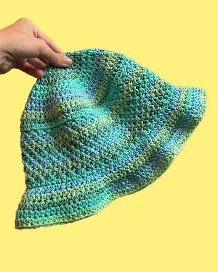 a hand is holding up a crocheted green and blue hat on a yellow background