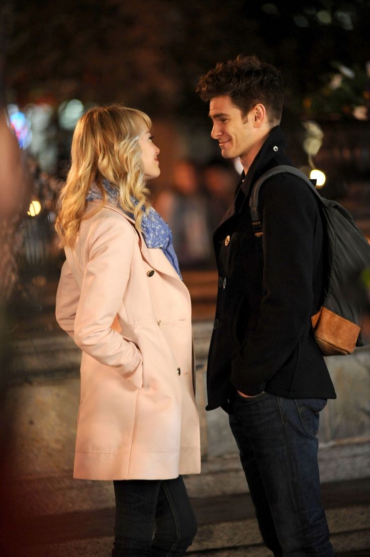 a man and woman standing next to each other on the street at night with city lights in the background